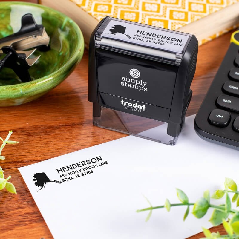 Simply Stamps branded Trodat 4913 self-inking return address stamp shown on a desk, with Alaska-themed address impression stamped on an envelope.