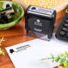 Simply Stamps branded Trodat 4913 self-inking return address stamp shown on a desk, with Alaska-themed address impression stamped on an envelope.
