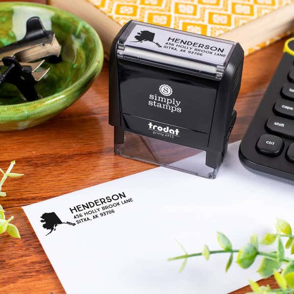 Simply Stamps branded Trodat 4913 self-inking return address stamp shown on a desk, with Alaska-themed address impression stamped on an envelope.