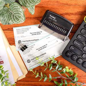 Trodat Printy 4916 self-inking stamp on desk next to printed checks, keyboard, and office supplies, showing use for endorsing or authorizing payments.
