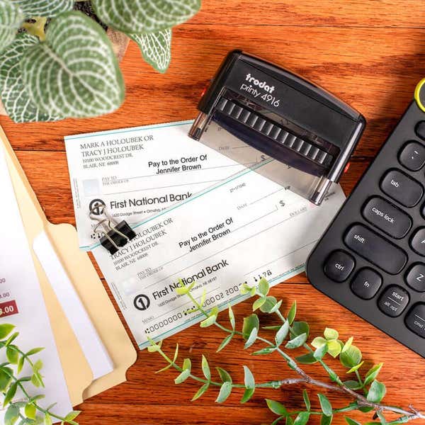 Trodat Printy 4916 self-inking stamp on desk next to printed checks, keyboard, and office supplies, showing use for endorsing or authorizing payments.
