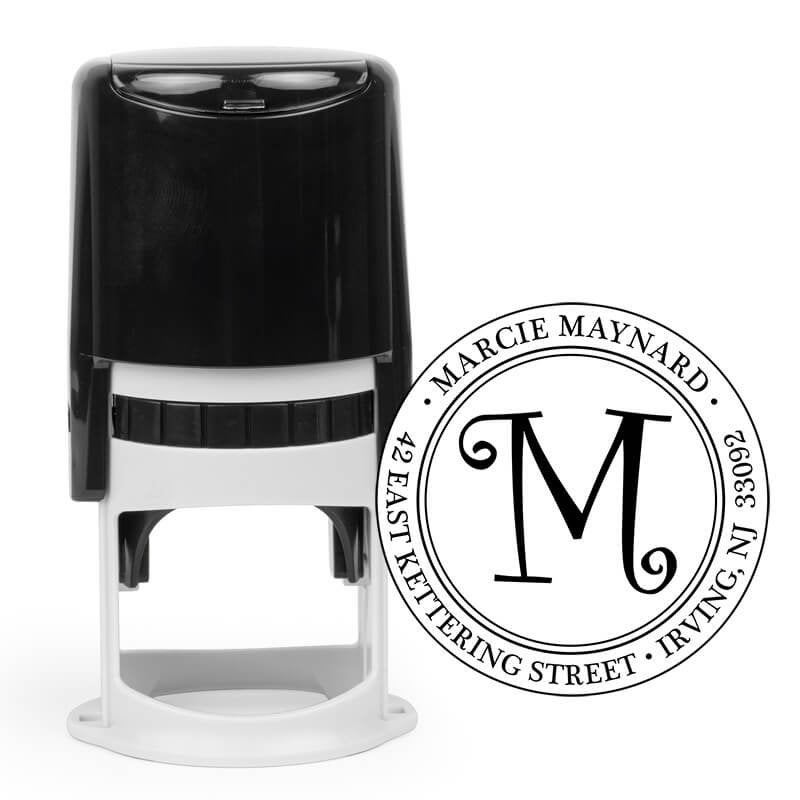 Round custom address stamp with ornate “M” design and return address, shown with self-inking stamp body.