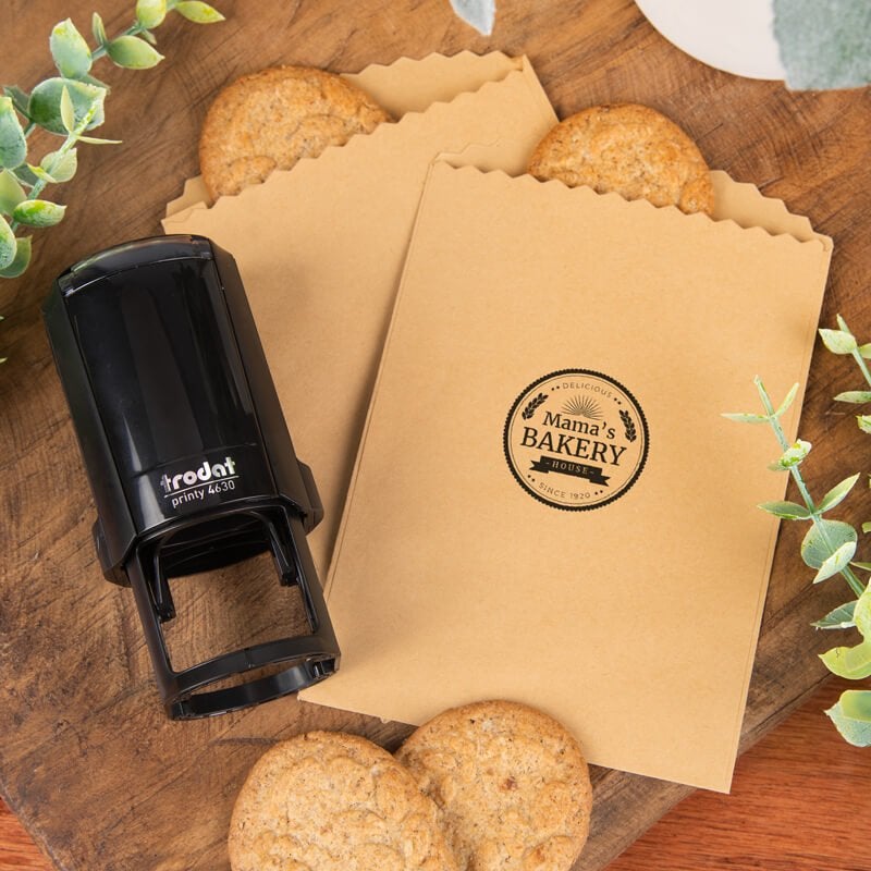 Custom round stamp impression applied to kraft paper treat bags, shown with a Trodat self-inking stamp