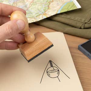 camp pot stamped in black on a cream notebook page beside a folded map, olive fabric and the wooden stamp in hand