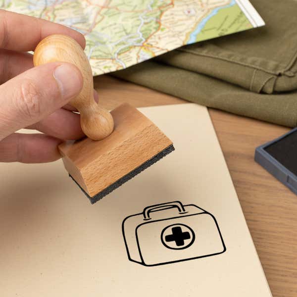 first aid kit stamped in black on a cream notebook page beside a folded map, olive fabric and the wooden stamp in hand