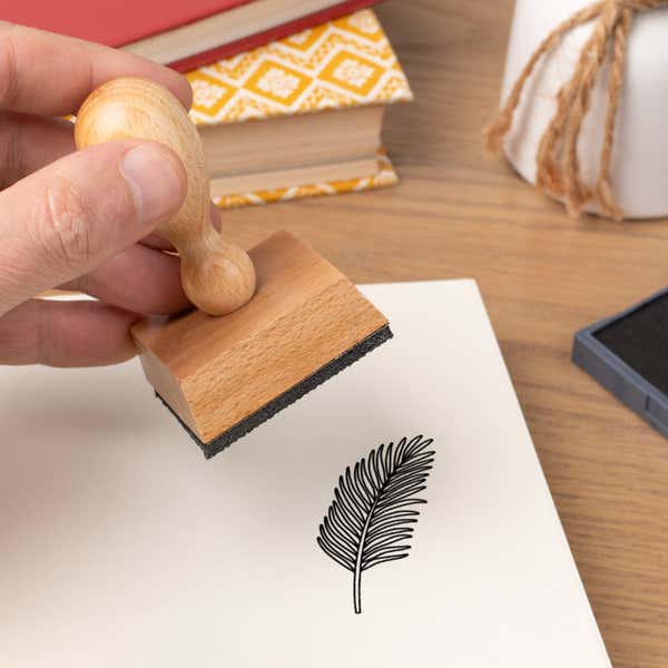 Hand pressing the Feathered Frond Craft Stamp onto cardstock beside a black ink pad