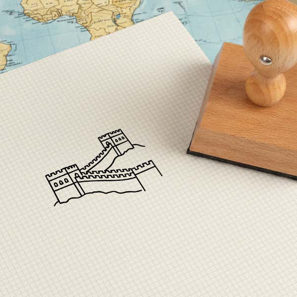 Great Wall of China stamped in black on cream graph paper laid over a world map, with the wooden stamp nearby