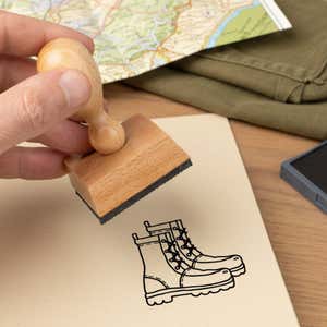 hiking boots stamped in black on a cream notebook page beside a folded map, olive fabric and the wooden stamp in hand