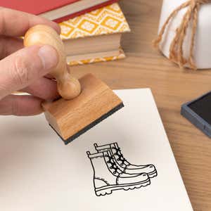Hand pressing the Hiking Boots Craft Stamp onto cardstock beside a black ink pad