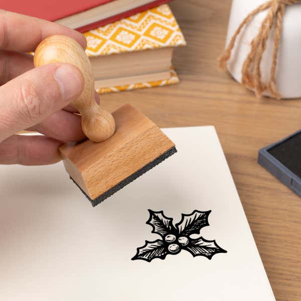 Hand pressing the Evergreen Holly Craft Stamp onto cardstock beside a black ink pad