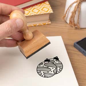 Hand pressing the Keepsake Tree Ornaments Craft Stamp onto cardstock beside a black ink pad