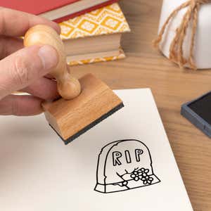 Hand pressing the Graveyard RIP Tombstone Craft Stamp onto cardstock beside a black ink pad