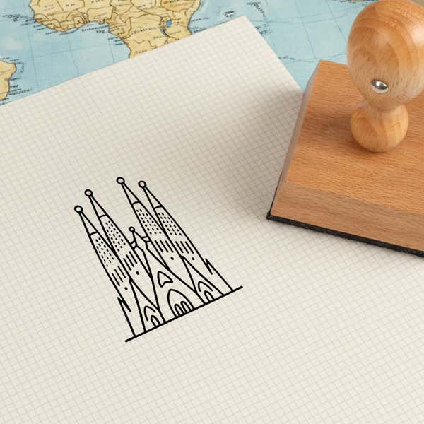 Sagrada Familia stamped in black on cream graph paper laid over a world map, with the wooden stamp nearby