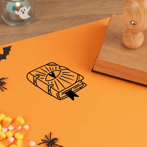 witchy spell book stamped in black on orange cardstock next to a wooden stamp, a paper bat cutout and candy corn