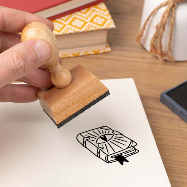 Hand pressing the Witchy Spell Book Craft Stamp onto cardstock beside a black ink pad