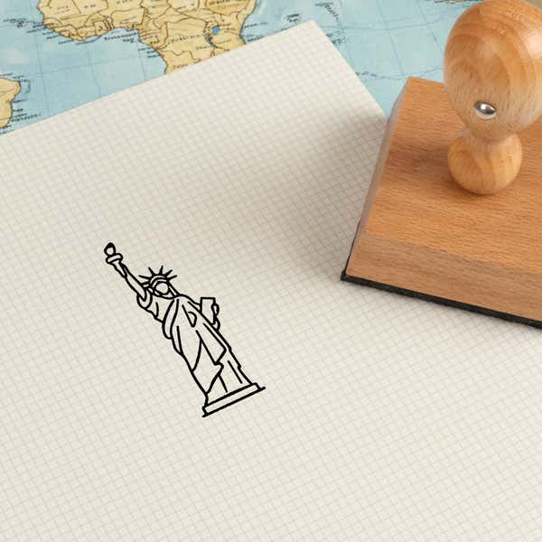 Statue of Liberty stamped in black on cream graph paper laid over a world map, with the wooden stamp nearby