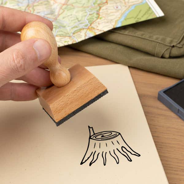 tree stump stamped in black on a cream notebook page beside a folded map, olive fabric and the wooden stamp in hand