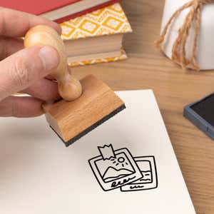 Hand pressing the Travel Photos Craft Stamp onto cardstock beside a black ink pad