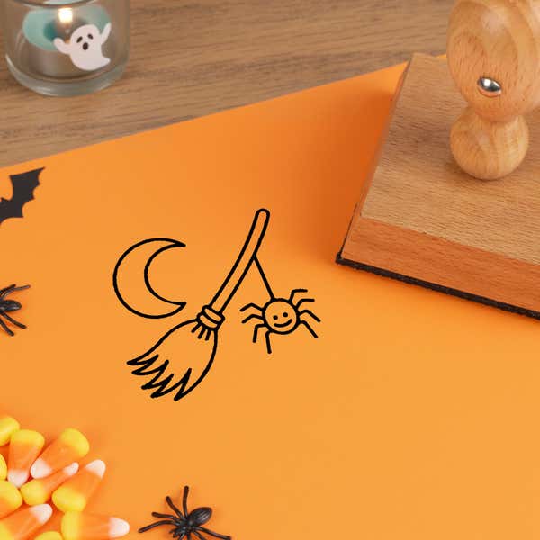 enchanted witch broom stamped in black on orange cardstock beside a wooden stamp, with plastic spiders and candy corn