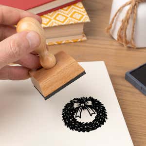 Hand pressing the Seasonal Christmas Wreath Craft Stamp onto cardstock beside a black ink pad