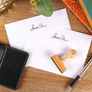 Santa Claus signature European hand stamp printed on the back flaps of two white envelopes with ink pad and stamp on wooden surface.
