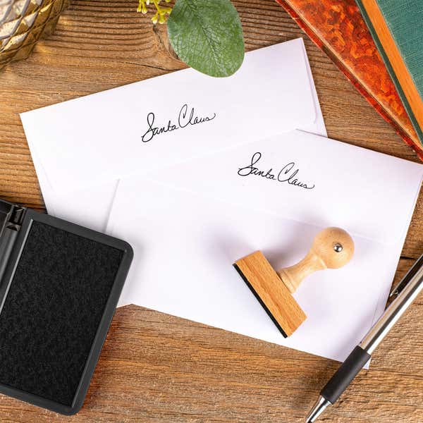 Santa Claus signature European hand stamp printed on the back flaps of two white envelopes with ink pad and stamp on wooden surface.