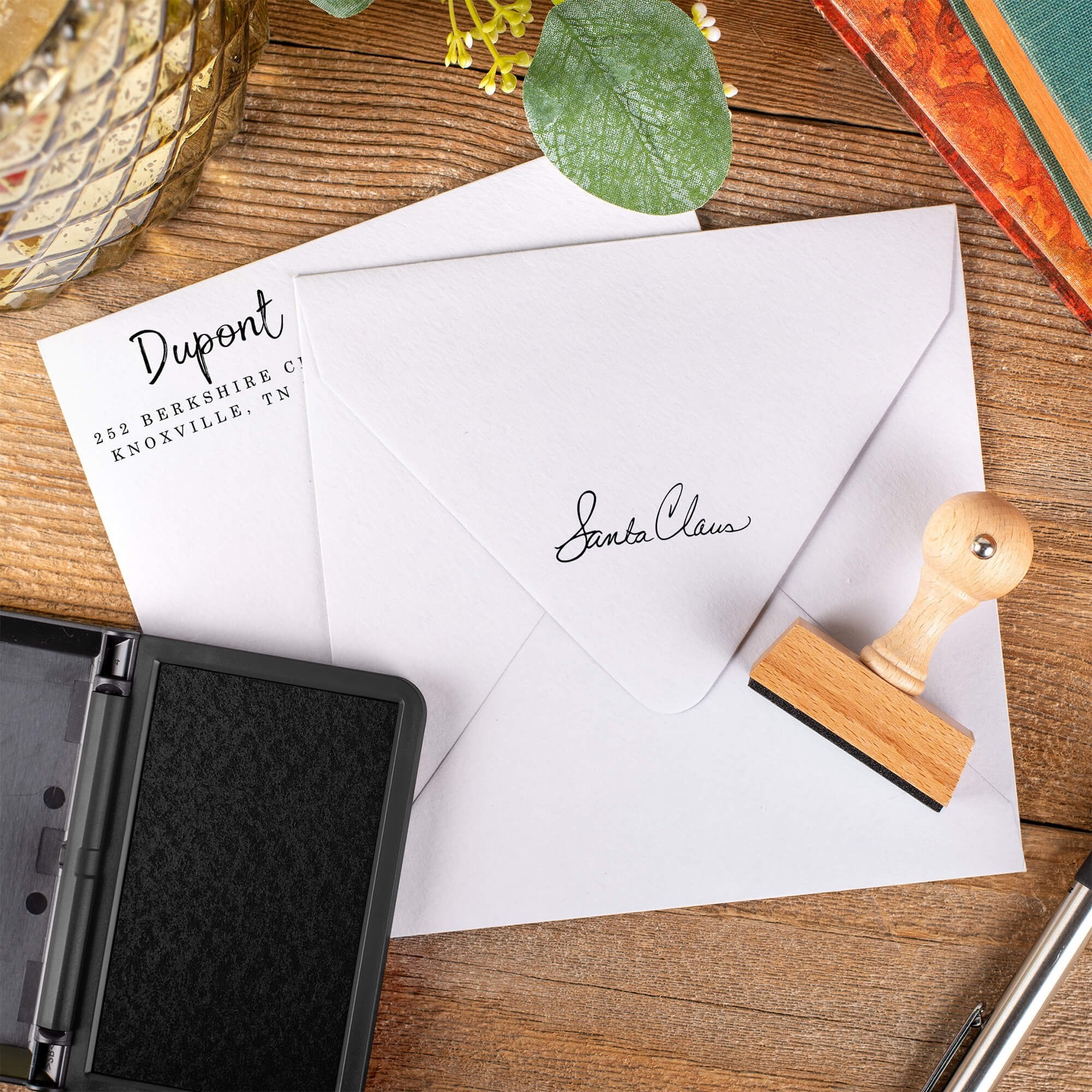 Santa Claus signature European hand stamp impression on front and back of two white envelopes on a wooden desk with ink pad and stamp.