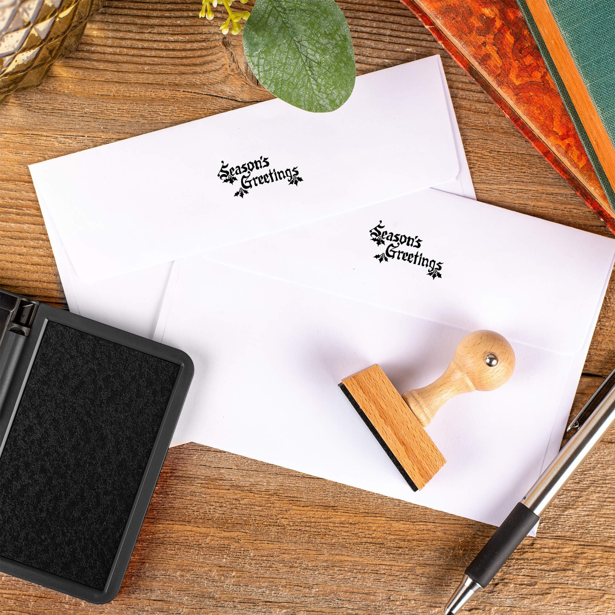 Season’s Greetings European hand stamp printed on the back flaps of two white envelopes with ink pad and wooden stamp on rustic wooden surface.