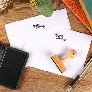 Season’s Greetings European hand stamp printed on the back flaps of two white envelopes with ink pad and wooden stamp on rustic wooden surface.