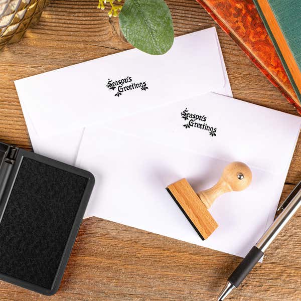 Season’s Greetings European hand stamp printed on the back flaps of two white envelopes with ink pad and wooden stamp on rustic wooden surface.