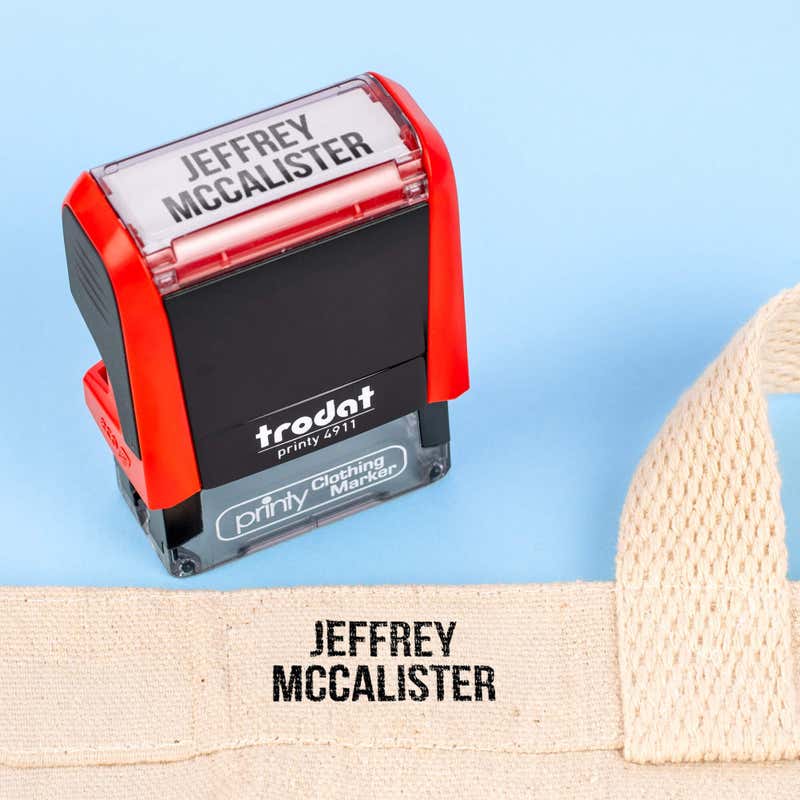 Natural canvas bag strap name-stamped in the Bold font, a heavy condensed all-caps print, next to a Trodat clothing stamp, on a blue background.