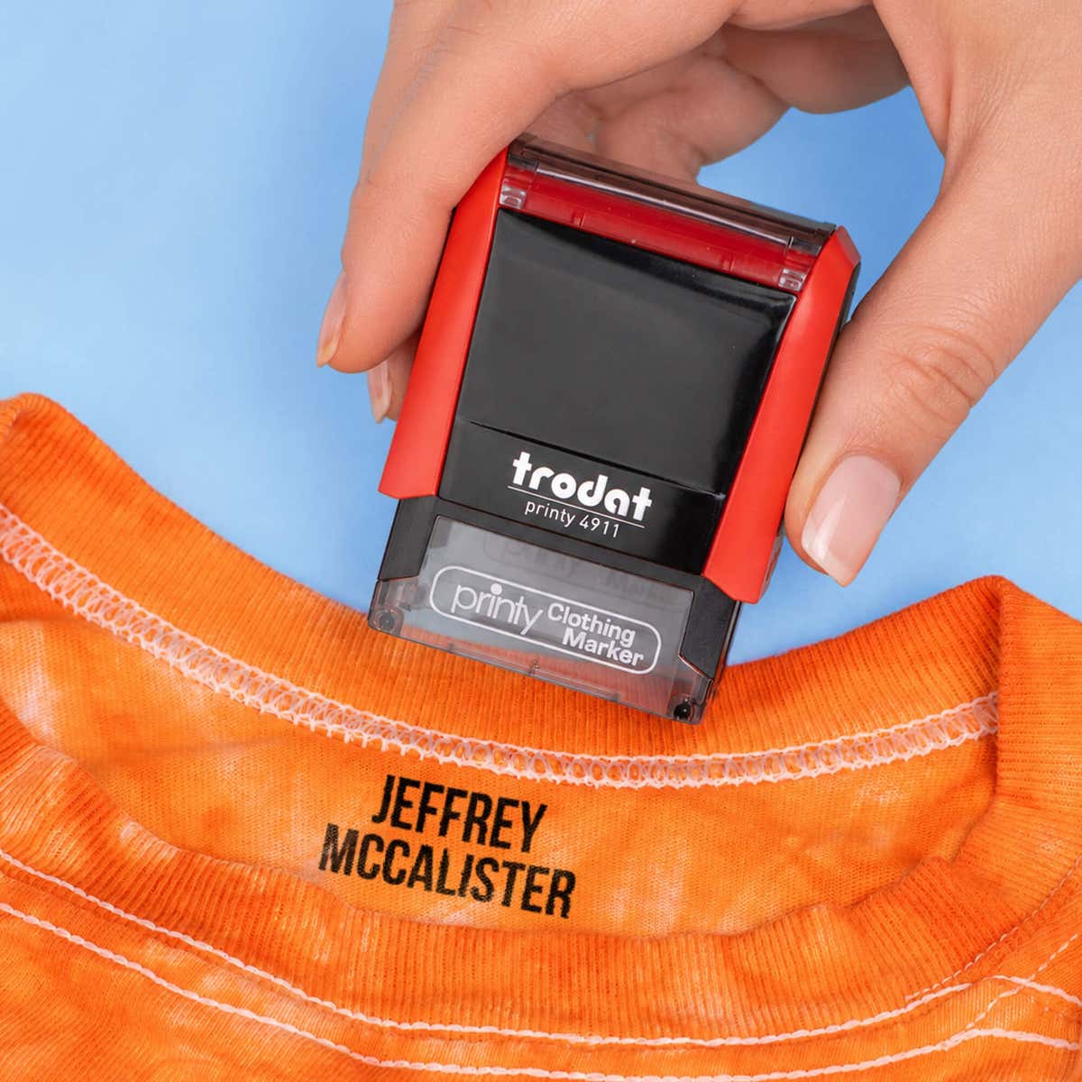 Hand stamping an orange t-shirt collar with a name in the Bold font, a heavy condensed all-caps print, using a Trodat clothing stamp.