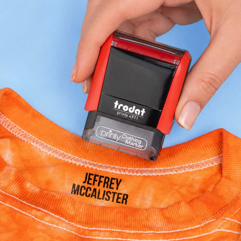 Hand stamping an orange t-shirt collar with a name in the Bold font, a heavy condensed all-caps print, using a Trodat clothing stamp.
