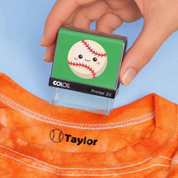Hand holding a baseball clothing name stamp above an orange shirt labeled Taylor