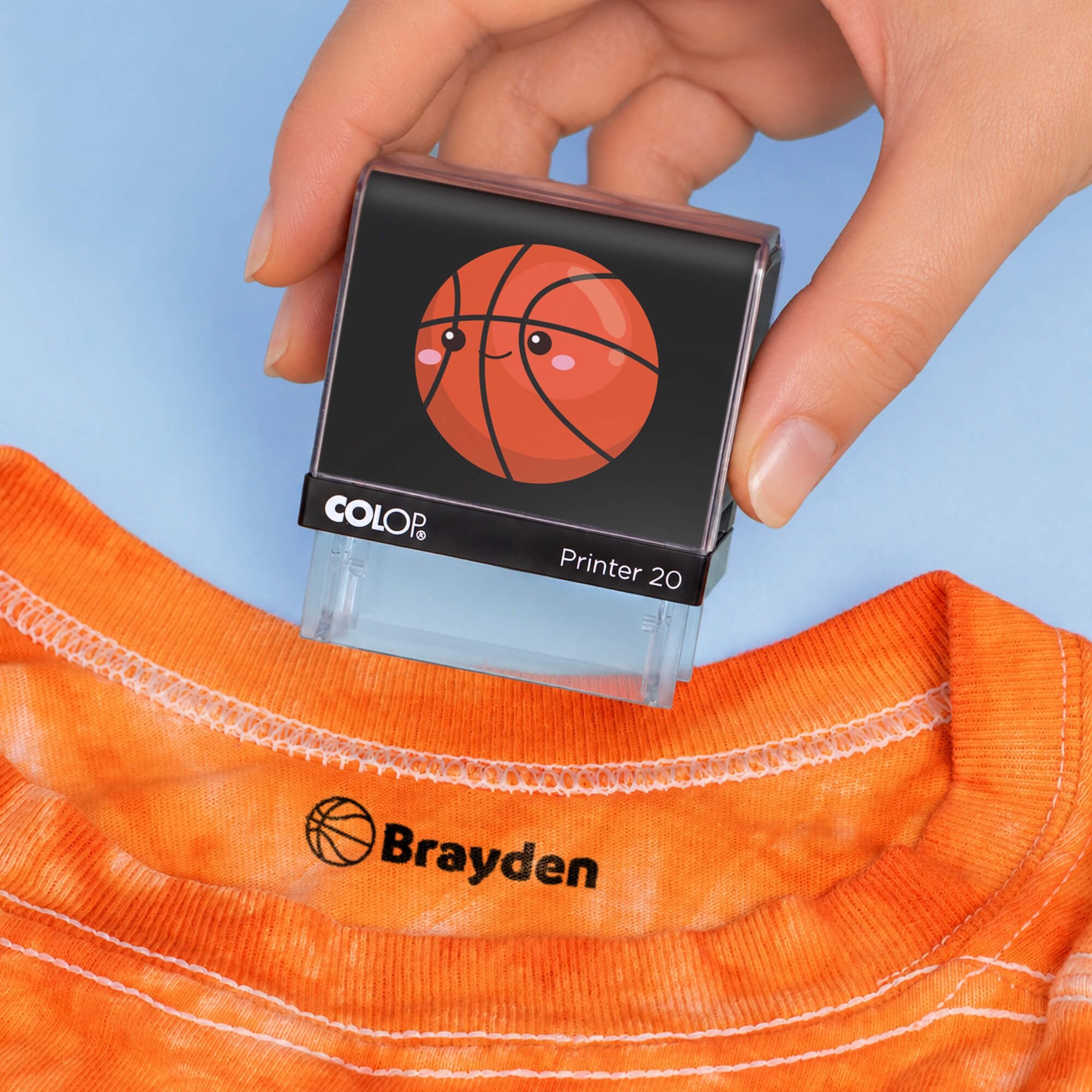 Hand holding a basketball clothing name stamp above an orange shirt labeled Brayden