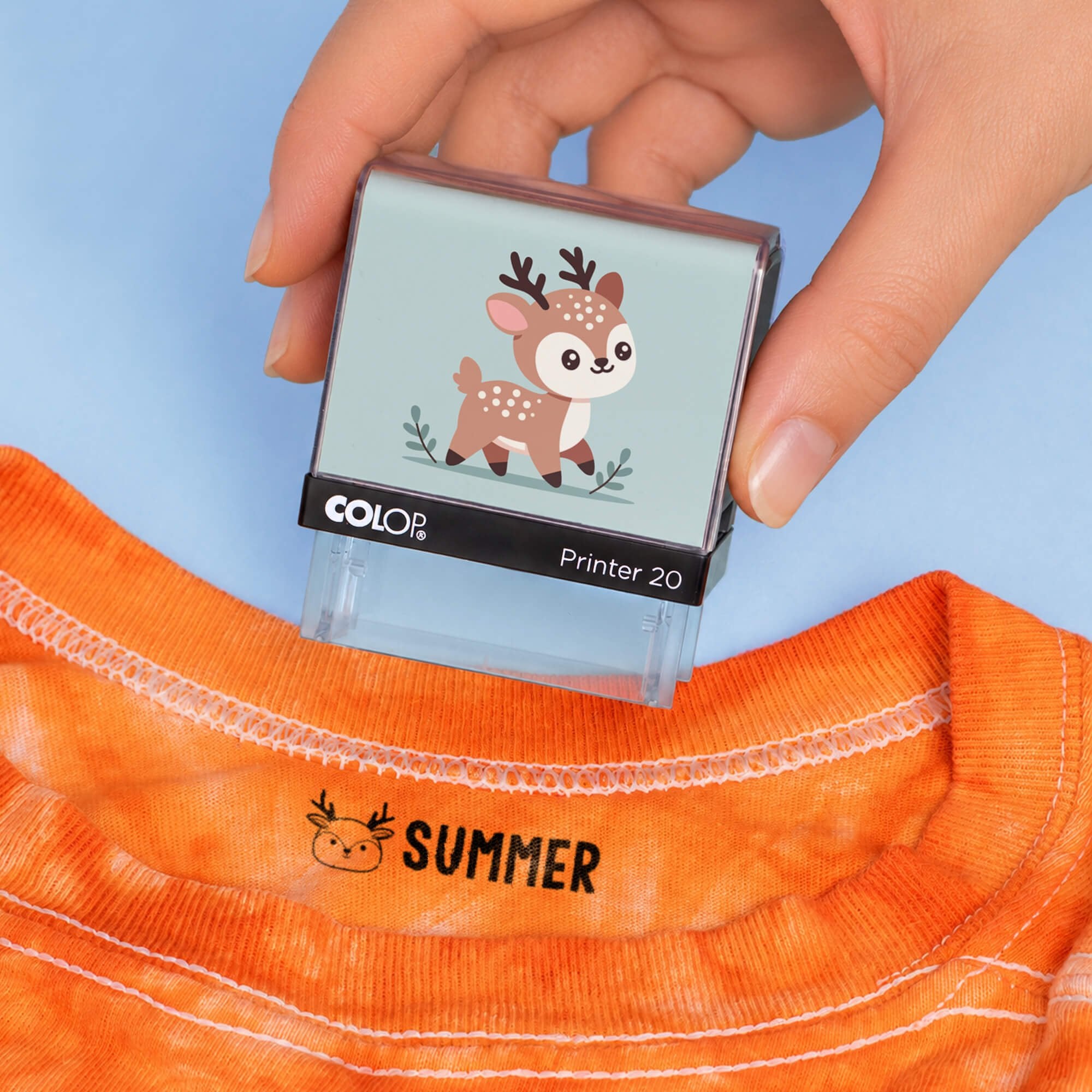 Hand holding a deer clothing name stamp above an orange shirt labeled Summer