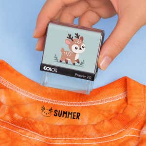 Hand holding a deer clothing name stamp above an orange shirt labeled Summer