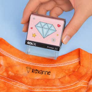 Hand holding a diamond clothing name stamp above an orange shirt labeled Roxanne