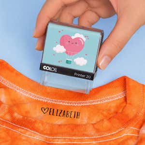 Hand holding a heart clothing name stamp above an orange shirt labeled Elizabeth