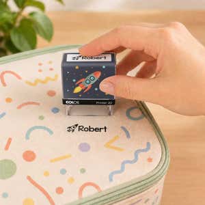 Hand using a rocket clothing name stamp to label a patterned lunch bag Robert