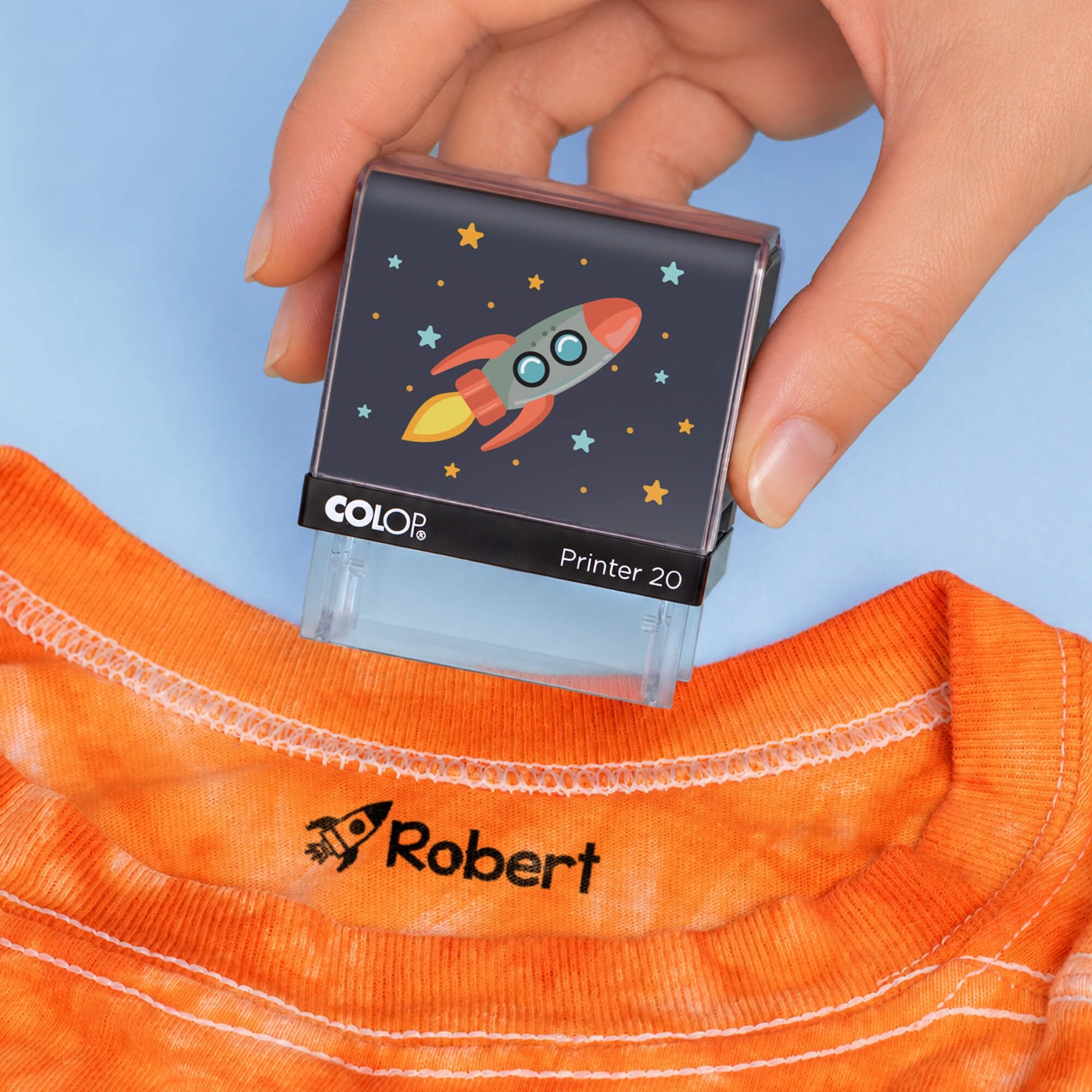 Hand holding a rocket clothing name stamp above an orange shirt labeled Robert