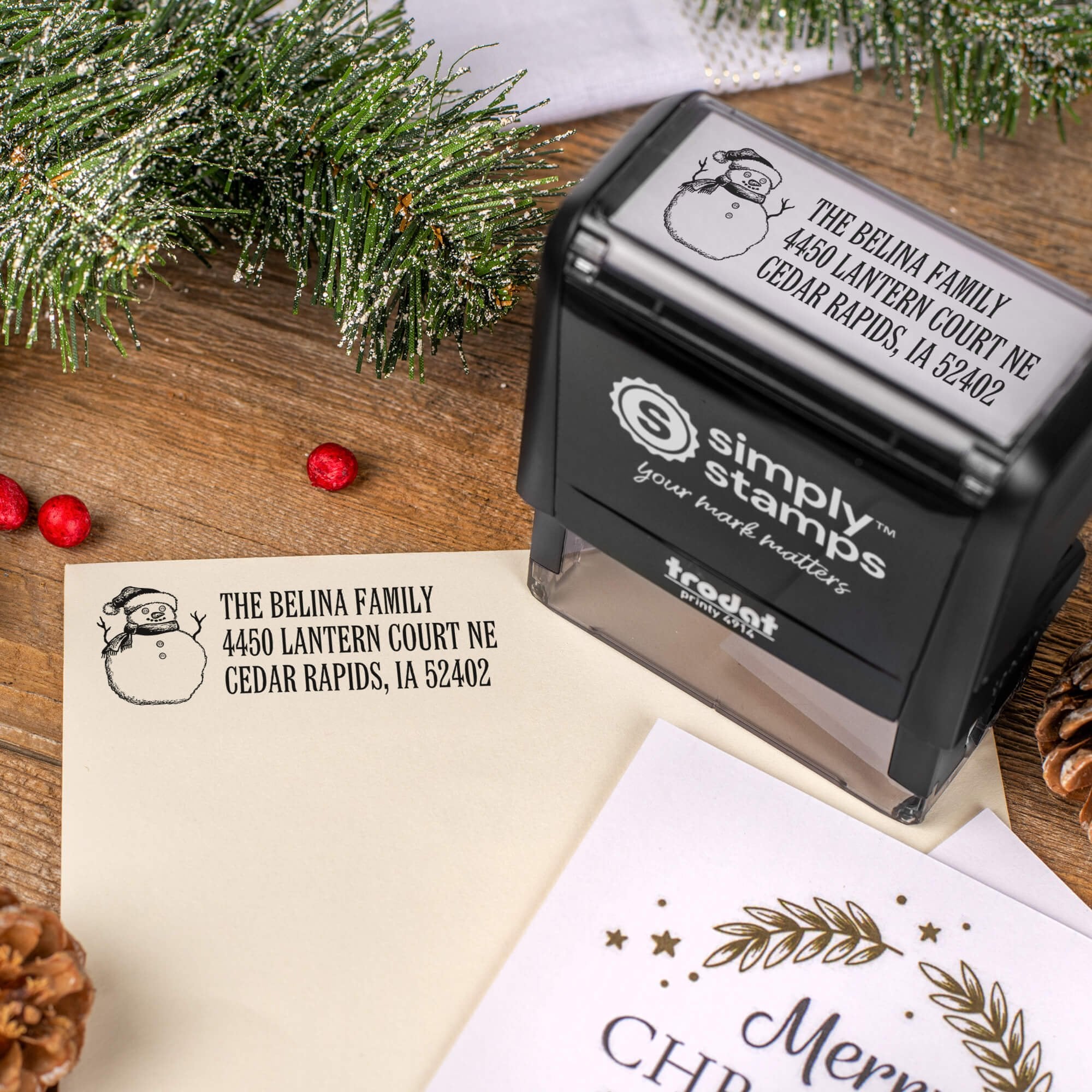 Holiday-themed ‘Sketch Snowman’ address stamp, hand using self-inking Christmas tree address stamp to personalize a holiday envelope on festive wooden table.