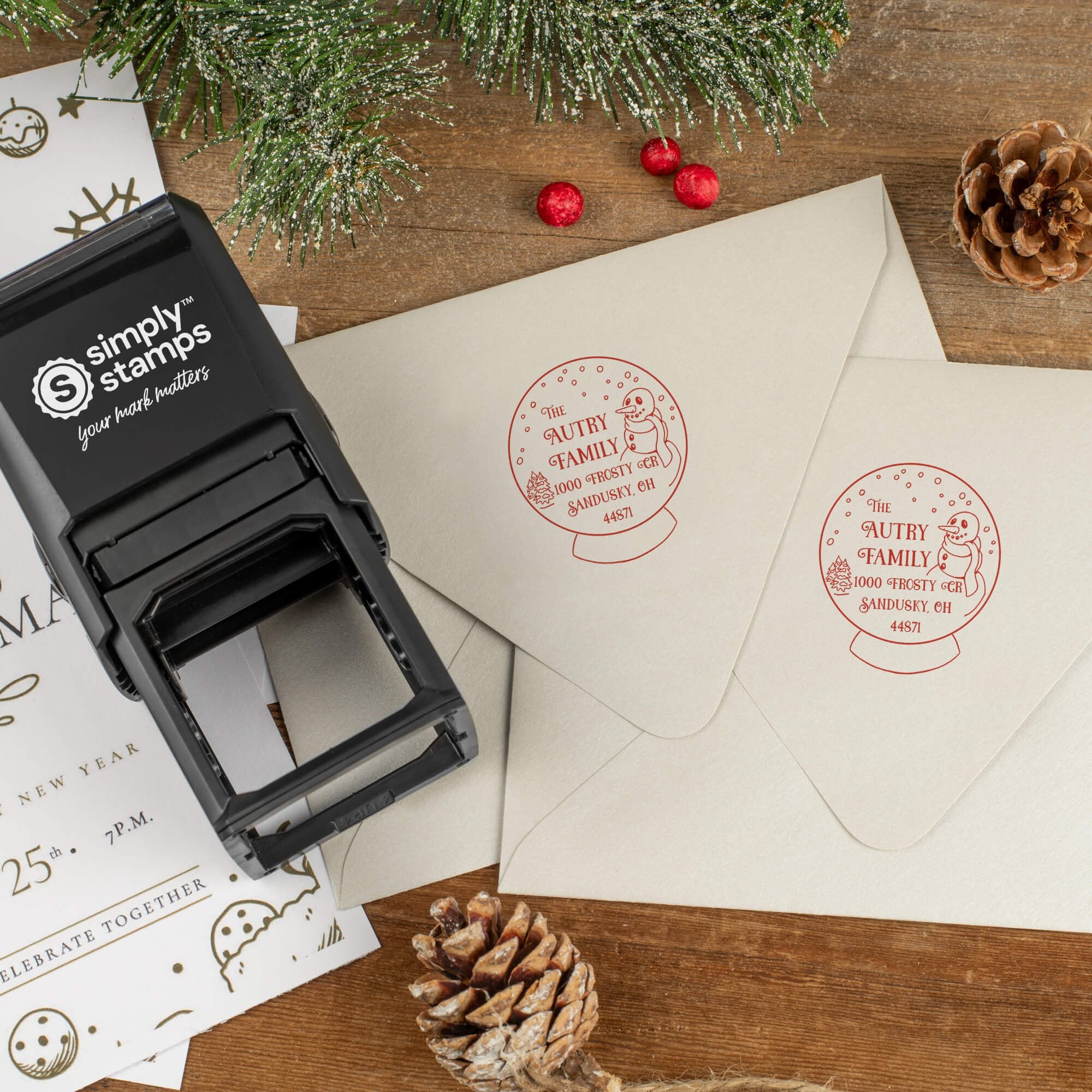 Holiday-themed ‘Snow Globe’ address stamp shown on envelopes beside festive stationery, pinecones, and decorations.