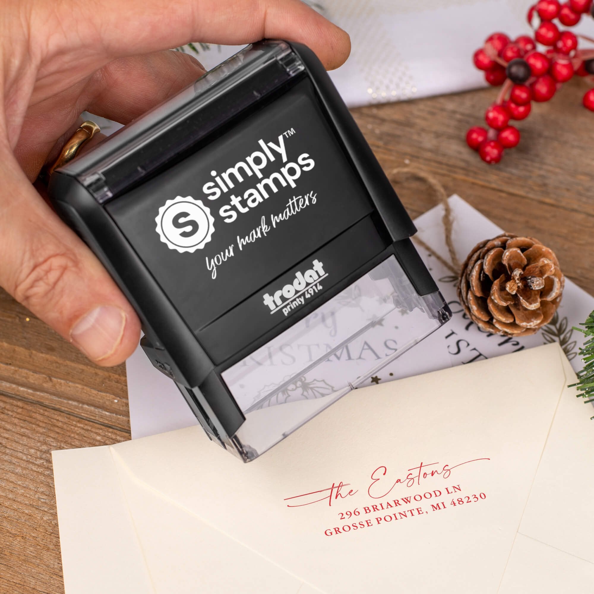 Holiday-themed ‘Signature Script Family Name’ address stamp, hand using self-inking Christmas tree address stamp to personalize a holiday envelope on festive wooden table.