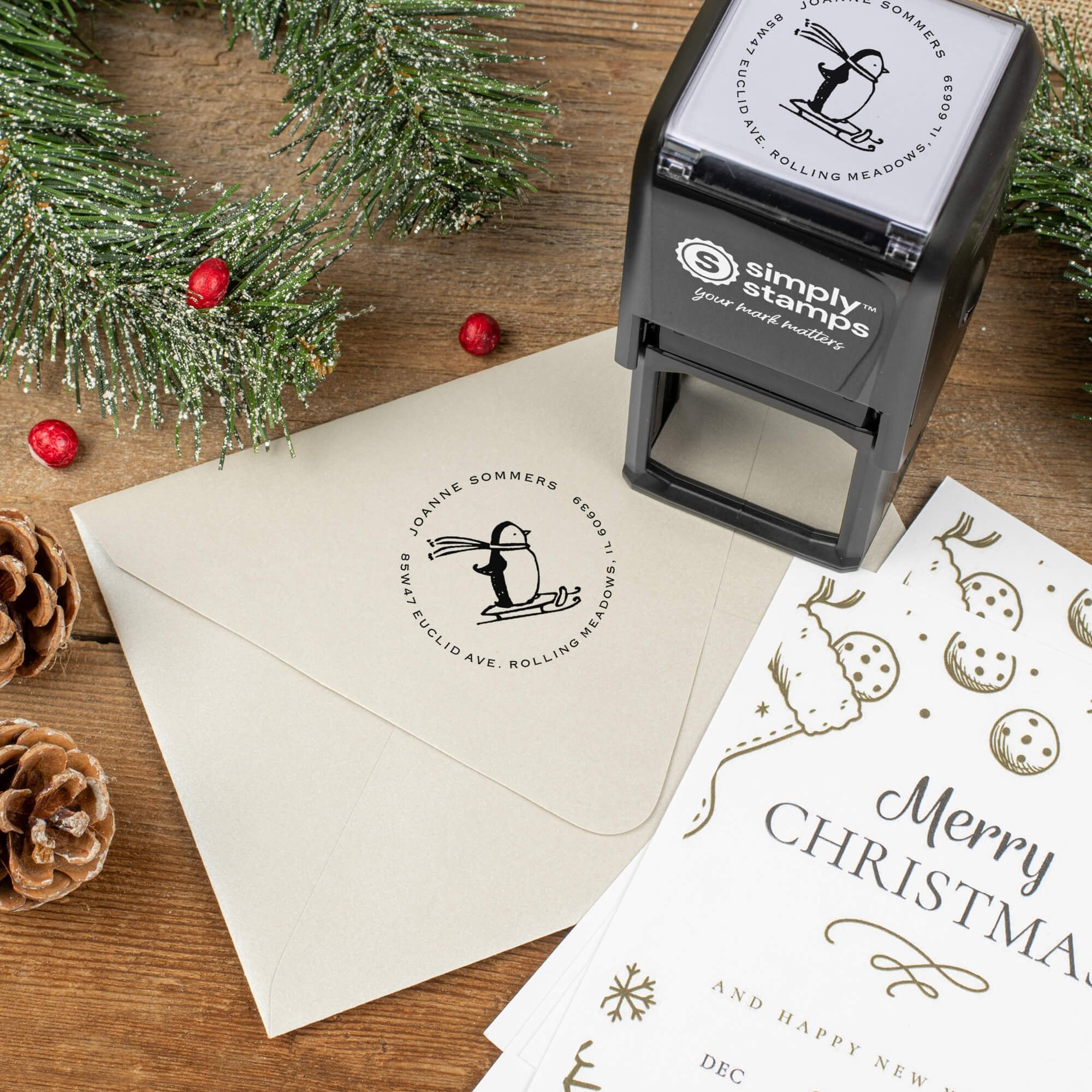 Holiday-themed ‘Sledding Penguin Holidaye’ address stamp shown on envelopes beside festive stationery, pinecones, and decorations.