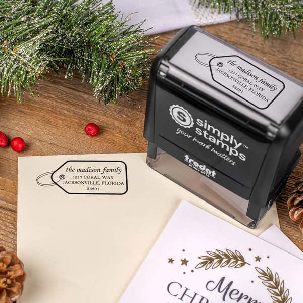 Holiday-themed ‘Madison Gift Tag’ address stamp, hand using self-inking Christmas tree address stamp to personalize a holiday envelope on festive wooden table.