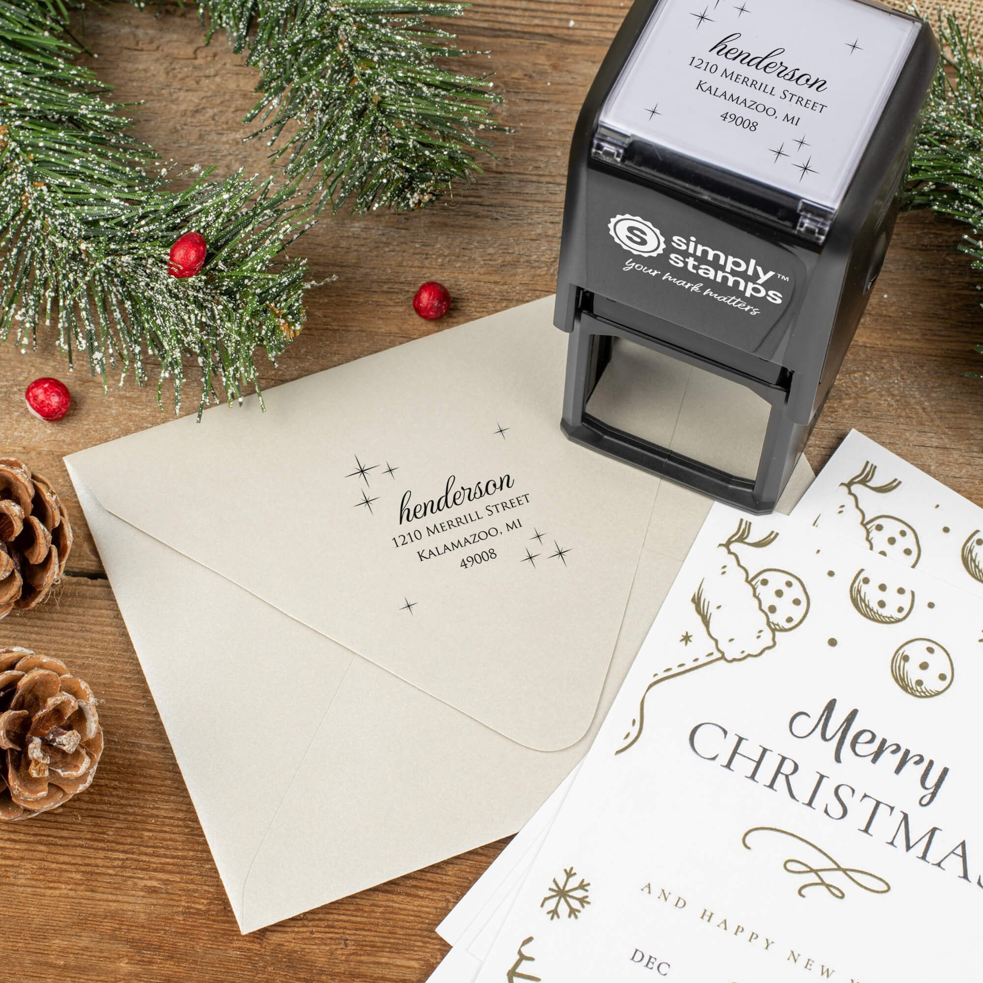 Holiday-themed ‘Henderson Twilight’ address stamp shown on envelopes beside festive stationery, pinecones, and decorations.
