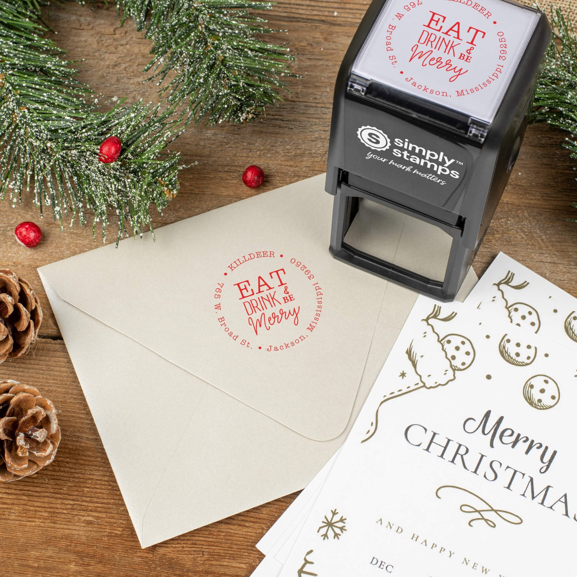 Holiday-themed ‘Eat Drink and Be Merry’ address stamp shown on envelopes beside festive stationery, pinecones, and decorations.
