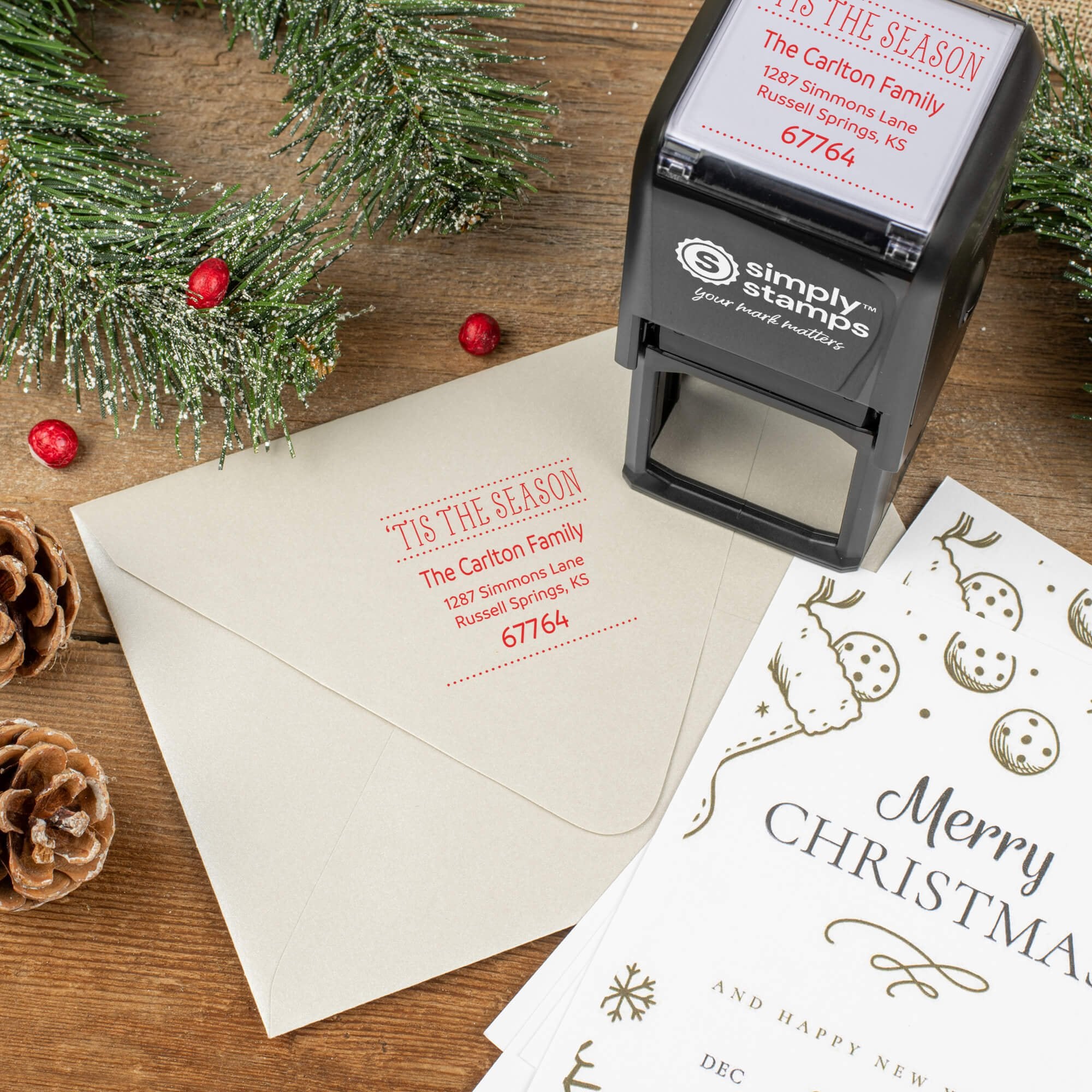 Holiday-themed ‘Tis The Season’ address stamp shown on envelopes beside festive stationery, pinecones, and decorations.