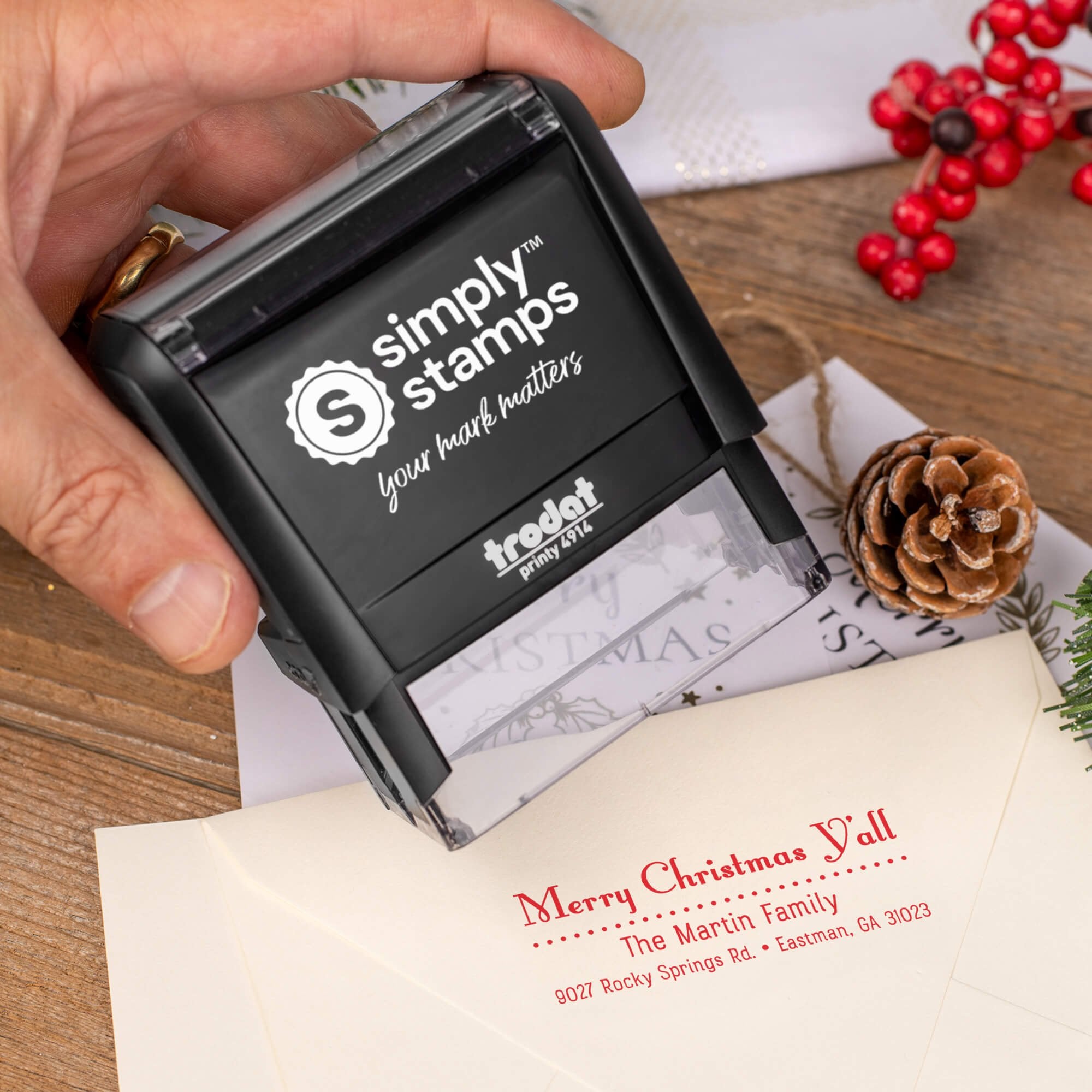 Holiday-themed ‘Merry Christmas Y’all’ address stamp, hand using self-inking Christmas tree address stamp to personalize a holiday envelope on festive wooden table.